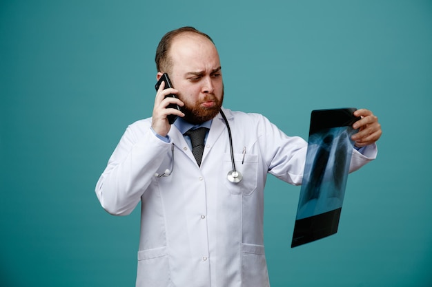 Ontevreden jonge mannelijke arts met een medische jas en stethoscoop om zijn nek die xray-opname vasthoudt en bekijkt terwijl hij aan de telefoon praat geïsoleerd op blauwe achtergrond