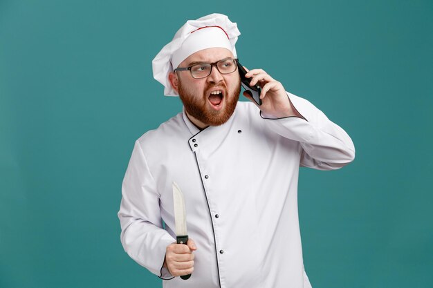 Ontevreden jonge mannelijke chef-kok met een uniforme bril en een pet met mes die naar de zijkant kijkt terwijl hij aan de telefoon praat geïsoleerd op blauwe achtergrond