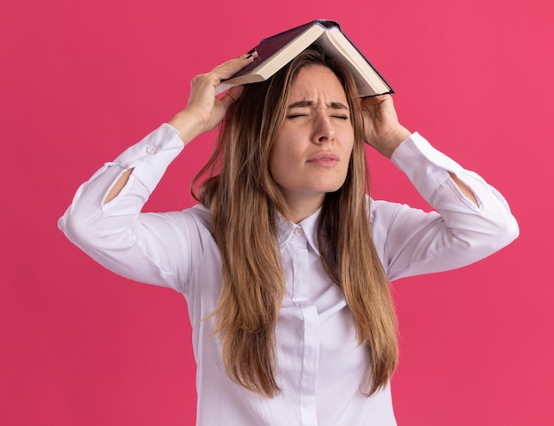 Ontevreden jonge, mooie blanke meid houdt een boek boven het hoofd geïsoleerd op een roze muur met kopieerruimte