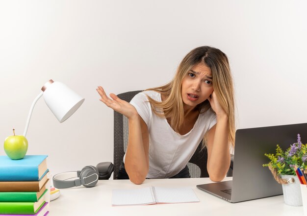 Ontevreden jonge mooie student meisje zit aan bureau met school tools hand op het hoofd zetten en lege hand tonen geïsoleerd op een witte achtergrond