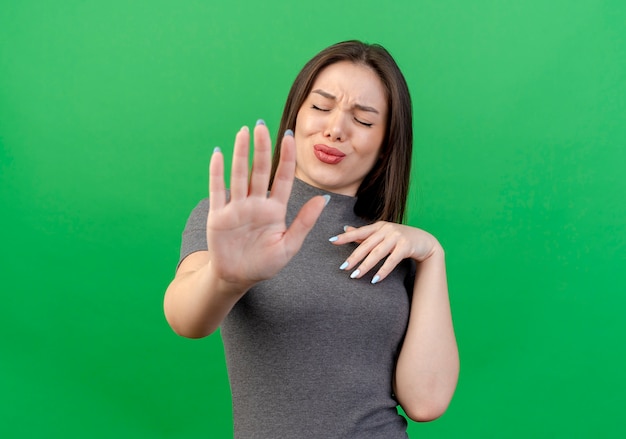 Ontevreden jonge mooie vrouw die hand op borst houdt en gebaren stop bij camera met gesloten ogen die op groene achtergrond met exemplaarruimte wordt geïsoleerd