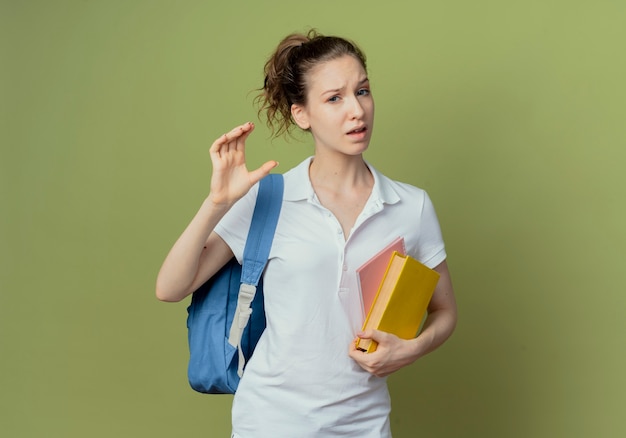 Ontevreden jonge mooie vrouwelijke student die het boek van de achterzakholding en notitieblok draagt die grootte toont die op olijfgroene achtergrond met exemplaarruimte wordt geïsoleerd