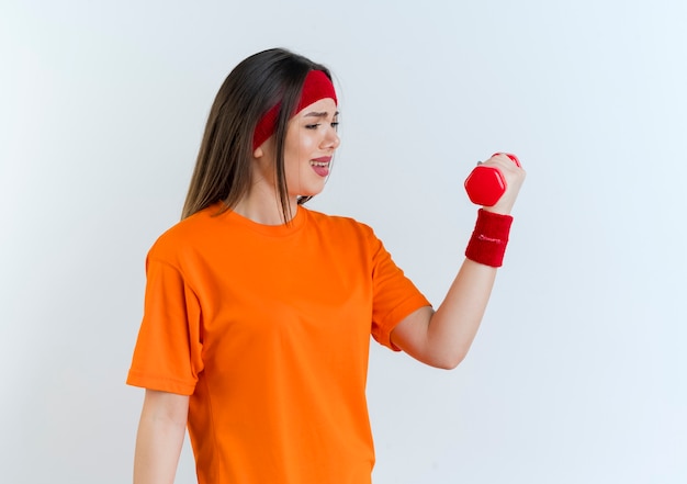 Ontevreden jonge sportieve vrouw die hoofdband en polsbandjes draagt die en domoor bekijken die op witte muur met exemplaarruimte wordt geïsoleerd