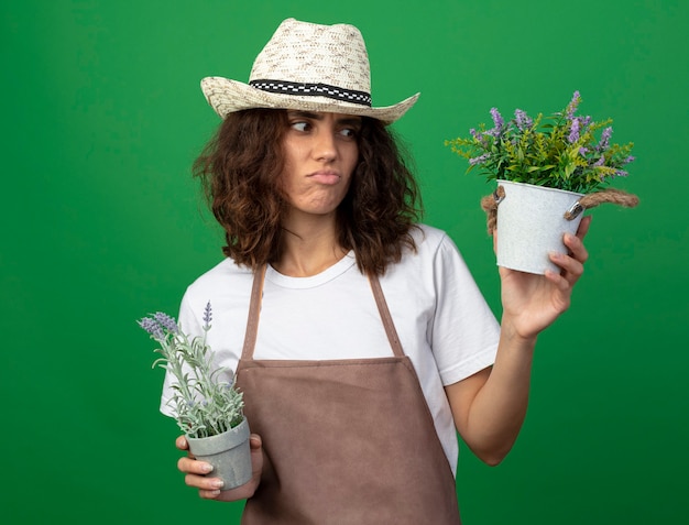 Gratis foto ontevreden jonge vrouw tuinman in uniform dragen tuinieren hoed bedrijf en kijken naar bloemen in bloempotten geïsoleerd op groen