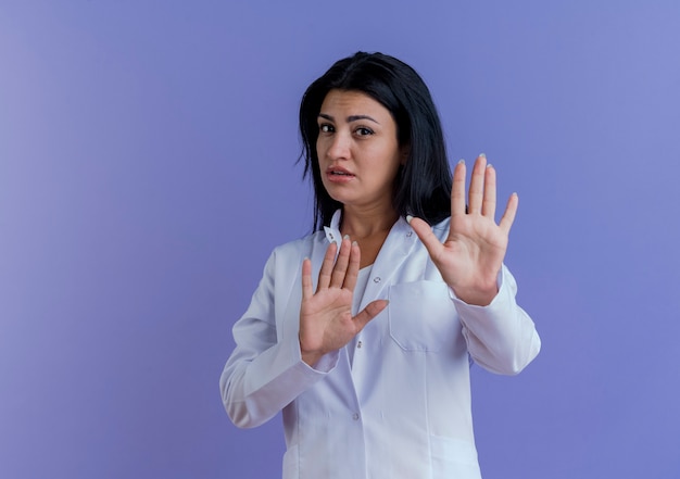 Ontevreden jonge vrouwelijke arts die medische mantel draagt die geen gebaar doet dat op purpere muur met exemplaarruimte wordt geïsoleerd