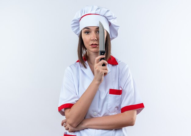 Ontevreden jonge vrouwelijke kok die chef-kok uniform bedekt oog draagt met mes geïsoleerd op een witte achtergrond