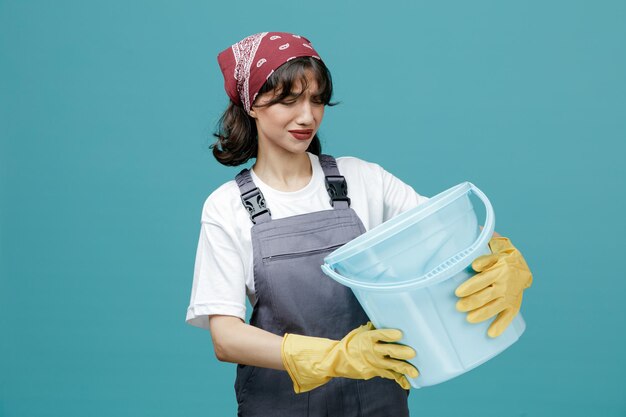 Ontevreden jonge vrouwelijke schoonmaker met een uniforme bandana en rubberen handschoenen die een emmer met beide handen vasthoudt en ernaar kijkt geïsoleerd op blauwe achtergrond