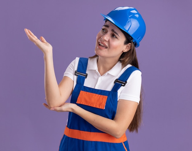 Ontevreden kijkend naar kant jonge bouwersvrouw in uniforme punten met hand aan kant die op purpere muur wordt geïsoleerd