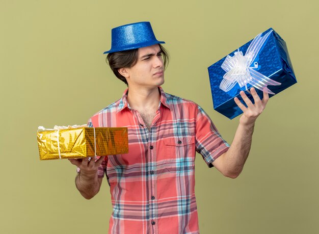 Ontevreden knappe blanke man met blauwe feesthoed houdt vast en kijkt naar geschenkdozen