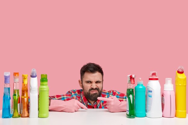 Ontevreden man met baard fronst gezicht van ontevredenheid, wijst naar schoonmaakspullen, draagt handschoenen, zit op desktop