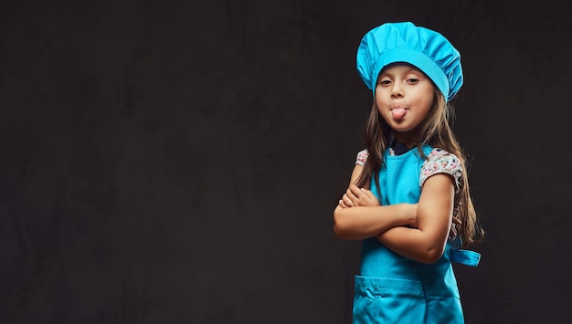 Ontevreden meisje gekleed in blauw koksuniform, staande met gekruiste armen en toont de uitgetrokken tong. Geïsoleerd op een donkere gestructureerde achtergrond.