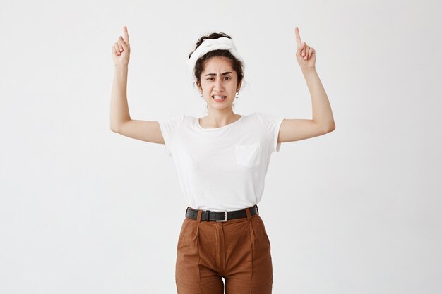 Ontevreden meisje met donker haar in knotje met vingers naar boven gericht, witte tanden op elkaar geklemd, wenkbrauwen fronsen. Jonge vrouw terloops gekleed met iets boven het hoofd, gebaren met wijsvingers