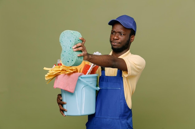 ontevreden met emmer schoonmaakgereedschap met spons jonge Afro-Amerikaanse schonere man in uniform met handschoenen geïsoleerd op groene achtergrond
