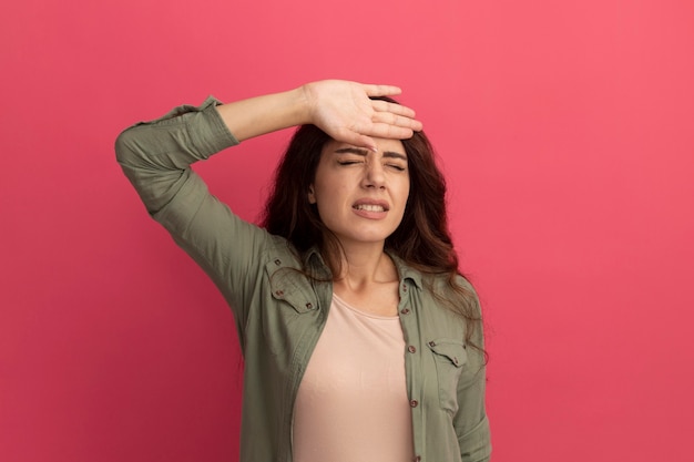 Ontevreden met gesloten ogen jong mooi meisje dat olijfgroen t-shirt draagt dat hand op voorhoofd zet dat op roze muur wordt geïsoleerd