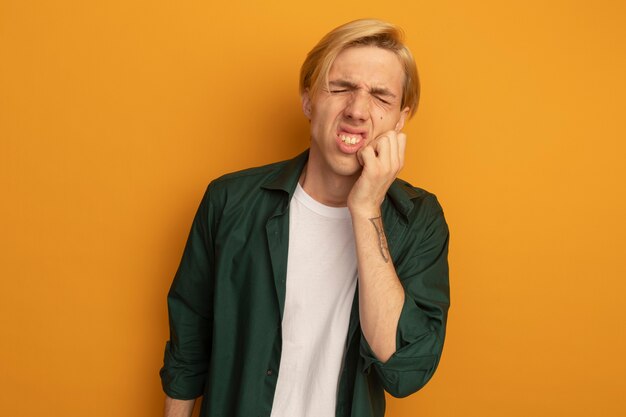 Ontevreden met gesloten ogen jonge blonde kerel die groen t-shirt draagt dat hand op wang zet