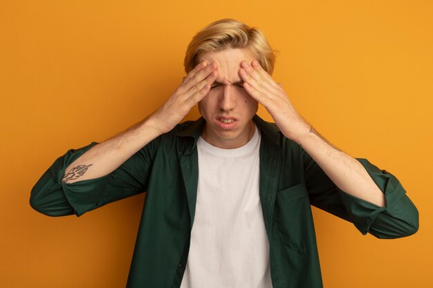 Ontevreden met gesloten ogen jonge blonde kerel die groen t-shirt draagt dat handen op voorhoofd legt