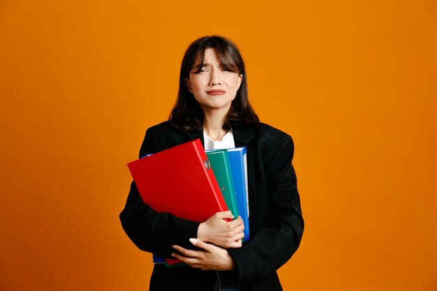 Ontevreden met mappen jonge mooie vrouw draagt zwarte jas geïsoleerd op een oranje achtergrond