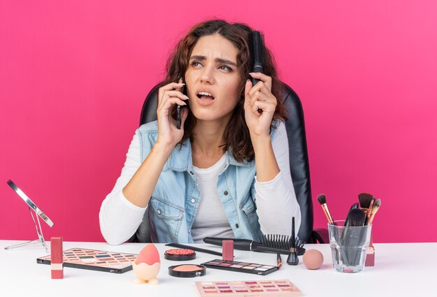 Ontevreden mooie blanke vrouw die aan tafel zit met make-uptools die aan de telefoon praat en haar haar kamt