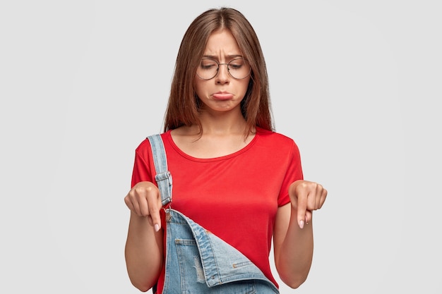 Ontevreden mooie brunette jonge vrouw met boos expressie, wijst naar beneden, merkt iets onaangenaams, heeft een boos uiterlijk, draagt modieuze overall met rood t-shirt, poseert tegen witte muur