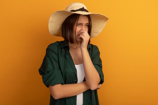 Ontevreden mooie vrouw met strandhoed sluit neus geïsoleerd op oranje muur