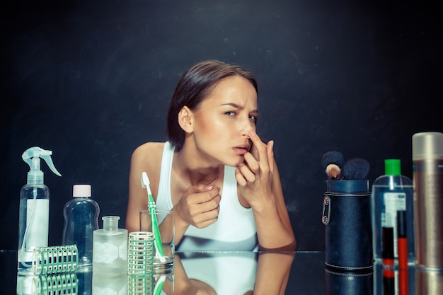 Ontevreden ongelukkige jonge vrouw die haar zelf in spiegel op zwarte studioachtergrond bekijkt. roblem huid en acne concept. Kaukasisch model in de studio