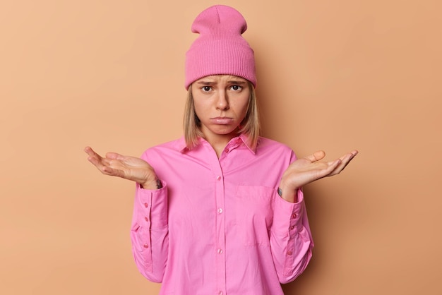 Ontevreden ongelukkige vrouw met blond haar ziet er helaas spreidt palmen uit heeft geen antwoord staat clueless gekleed in casual roze shirt en hoed poses tegen bruine achtergrond. Twijfel en aarzeling concept