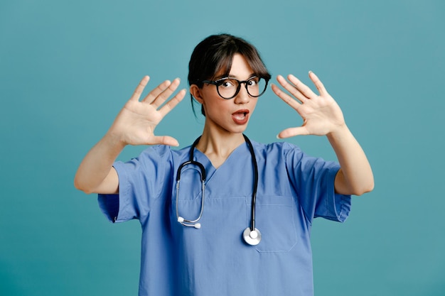 Ontevreden opgeheven hand jonge vrouwelijke arts die uniforme stethoscoop draagt die op blauwe achtergrond wordt geïsoleerd