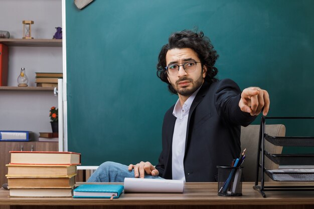 Ontevreden punten aan de voorkant van de mannelijke leraar die een bril draagt die aan tafel zit met schoolhulpmiddelen in de klas