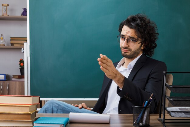 Ontevreden punten aan de zijkant van een mannelijke leraar die een bril draagt die aan tafel zit met schoolgereedschap in de klas