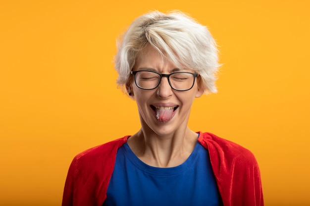 Gratis foto ontevreden supervrouw met rode cape in optische glazen steekt tong uit die op oranje muur wordt geïsoleerd