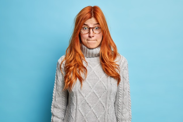 Ontevreden verbaasde roodharige jonge vrouw drukt lippen en kijkt beschaamd heeft geschokte uitdrukking gekleed in trui en bril.