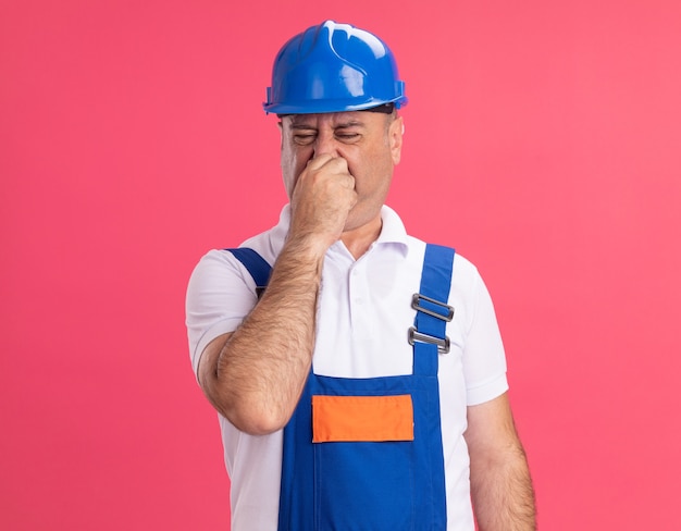 Ontevreden volwassen bouwersmens in uniform houdt neus vast die op roze muur wordt geïsoleerd