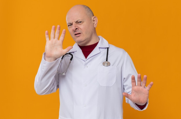 Ontevreden volwassen slavische man in doktersuniform met stethoscoop die handen open houdt en stoptekent