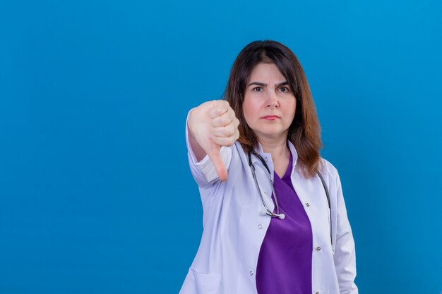 Ontevreden vrouw arts van middelbare leeftijd, gekleed in witte jas en met een stethoscoop met fronsend gezicht met duim omlaag staande over blauwe ruimte