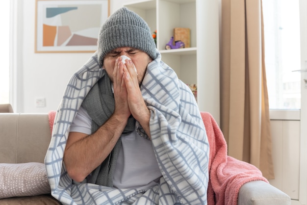 Ontevreden zieke slavische man met sjaal om nek met wintermuts gewikkeld in plaid veegt zijn neus af met tissue zittend op de bank in de woonkamer