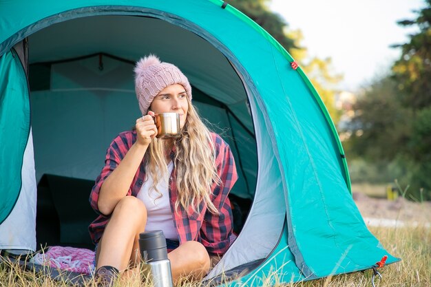 Ontspannen blonde vrouw in hoed die thee drinkt, in tent zit en weg kijkt. Kaukasische langharige vrouwelijke toeristenzitting op gazon. Toerisme, avontuur en zomervakantie concept