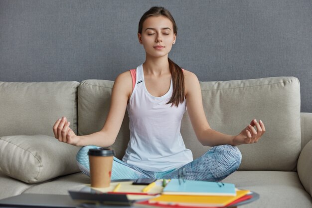 Ontspannen geconcentreerde tiener gekleed in vrijetijdskleding, zit in yogapositie, mediteert en heeft pauze na hard werken, drinkt afhaalkoffie