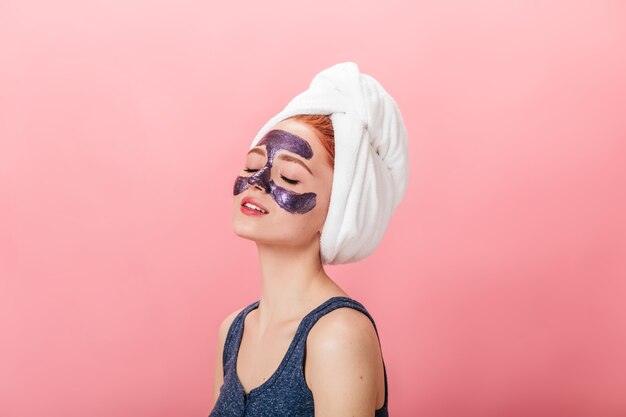 Ontspannen jonge vrouw die kuur op roze achtergrond doet. Studio shot van blij meisje met gezichtsmasker poseren met gesloten ogen.