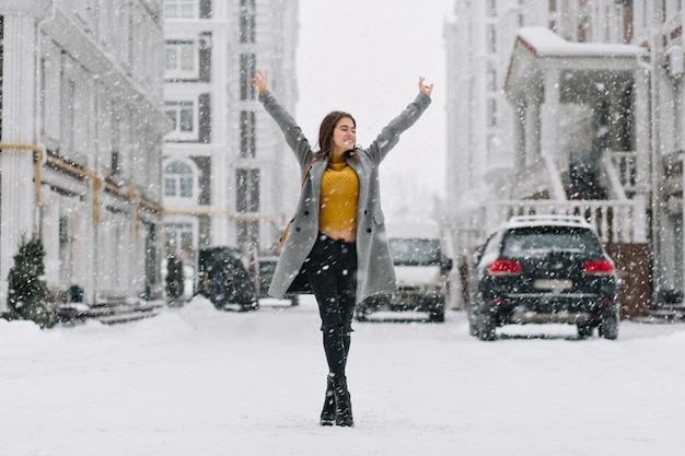 Ontspannen Kaukasische vrouw poseren met handen omhoog onder sneeuwval op stedelijke straat. Buiten foto van gemiddelde lengte van mooie vrouw in gele trui en grijze jas genieten van weekend in winter stad.