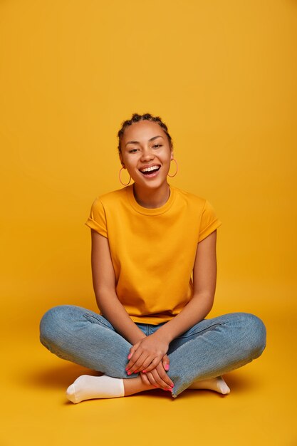 Ontspannen mooie duizendjarige vrouw zit gekruiste benen, lacht, heeft een hilarisch gesprek, draagt t-shirt, spijkerbroek, sokken, heeft cornrow-kapsel, geïsoleerd op gele muur, drukt zorgeloze emoties uit