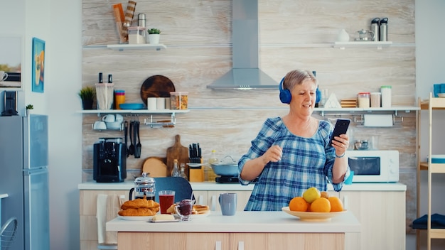 Ontspannen senior vrouw luisteren muziek op koptelefoon tijdens het ontbijt in de keuken. Ouderen dansen, leuke levensstijl met moderne technologie