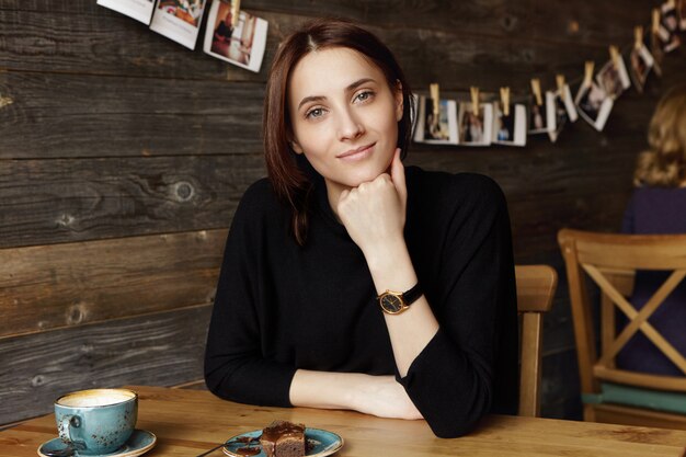 Ontspannen vertrouwen jonge vrouw met donker haar, gekleed in zwarte kleding kijken met charmante glimlach, elleboog rusten op houten tafel met mok en dessert terwijl het hebben van koffie in restaurant