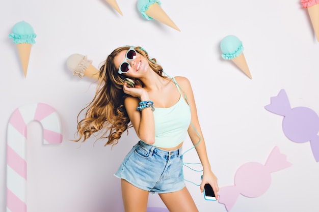Ontspannend meisje met lange glanzende krullen dansen in haar kamer met pastel interieur mobiele telefoon in de hand houden. portret van huiveringwekkende jonge vrouw met een lichtblauwe tanktop en een zonnebril.