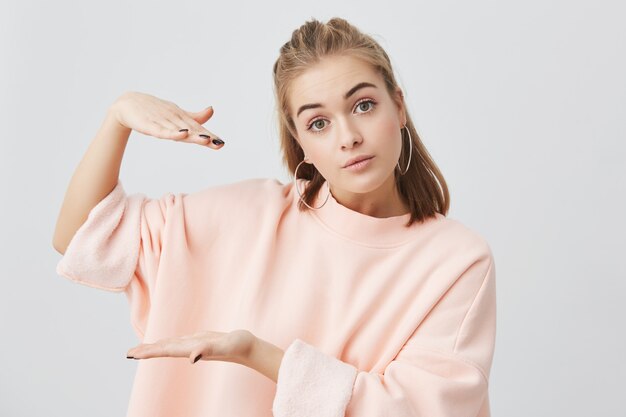 Ontsproten van leuk charmant meisje dat modieus roze sweatshirt met lange mouwen draagt dat de grootte van iets met handen toont, actief gesturing. Blonde Europese vrouw poseren.