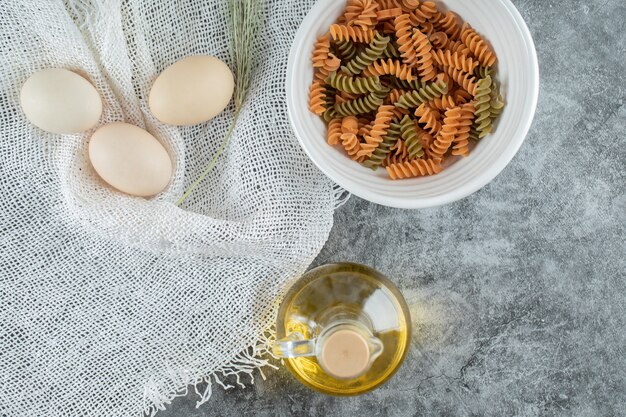 Onvoorbereide spiraalvormige macaroni in witte plaat met drie eieren en een fles olie