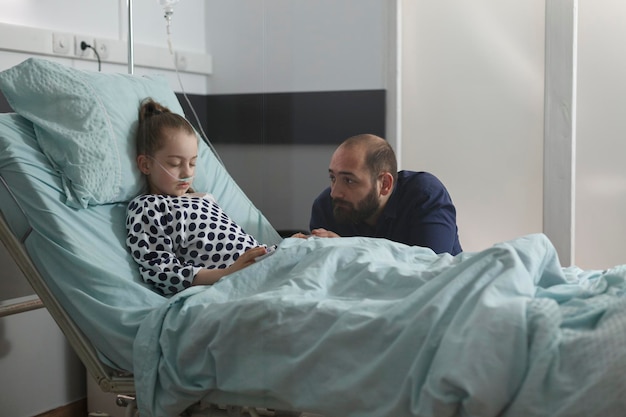 Onwel klein kind met een neusslang die rust terwijl de ouder wacht om wakker te worden uit de anesthesie. Trieste vader zit naast gehospitaliseerd ziek meisje rustend op het bed van de patiënt in de kinderafdeling van het ziekenhuis.