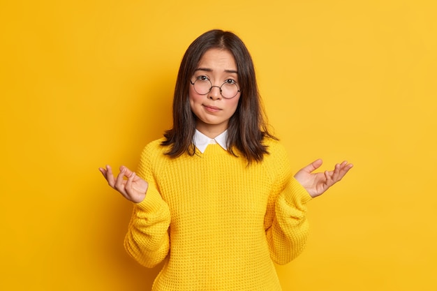 Onwetende aarzelende Aziatische vrouw haalt schouders op in een onnozel gebaar en kan geen beslissing nemen, draagt een ronde bril en een losse trui.