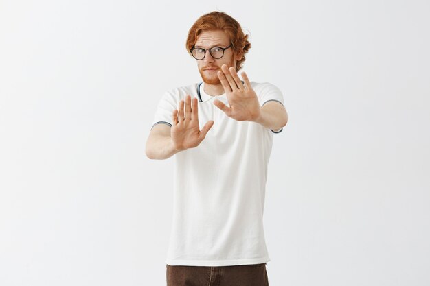 Gratis foto onwillige bebaarde roodharige man poseren tegen de witte muur met bril