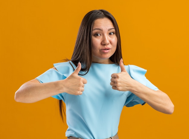 Onzeker jong Aziatisch meisje dat naar de camera kijkt en duimen omhoog houdt geïsoleerd op een oranje muur met kopieerruimte