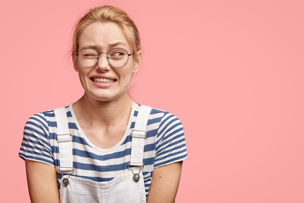 Gratis foto onzekere blonde vrouw houdt één oog dicht, kijkt opzij met een bange uitdrukking, geschrokken om iets onaangenaams te horen